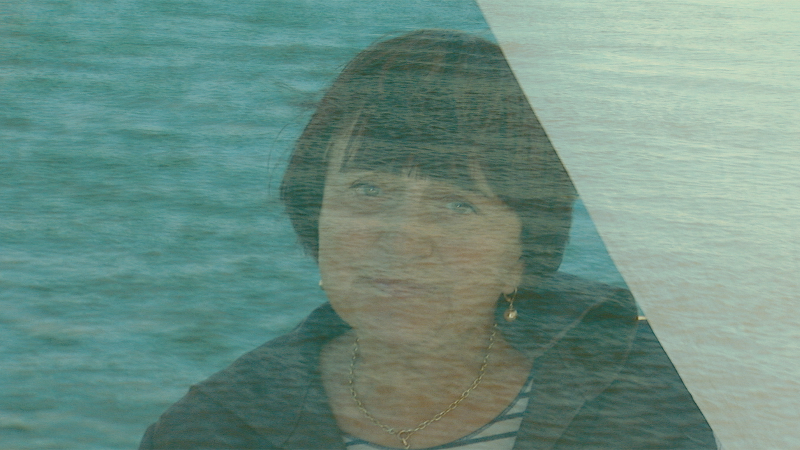 In this still from Agnès Varda's 2008 film, "The Beaches of Agnès," a close-up of Agnès's head and shoulders shows her looking toward the camera through a visual overlay that looks like a watery film and gives the image a bluish cast. As a result of the special effect, she looks slightly spectral. She has a wistful expression on her face. She wears a bowl cut, dangly gold earrings, and a necklace. The collar of her jacket is turned up on one side, as if whipped by the wind.