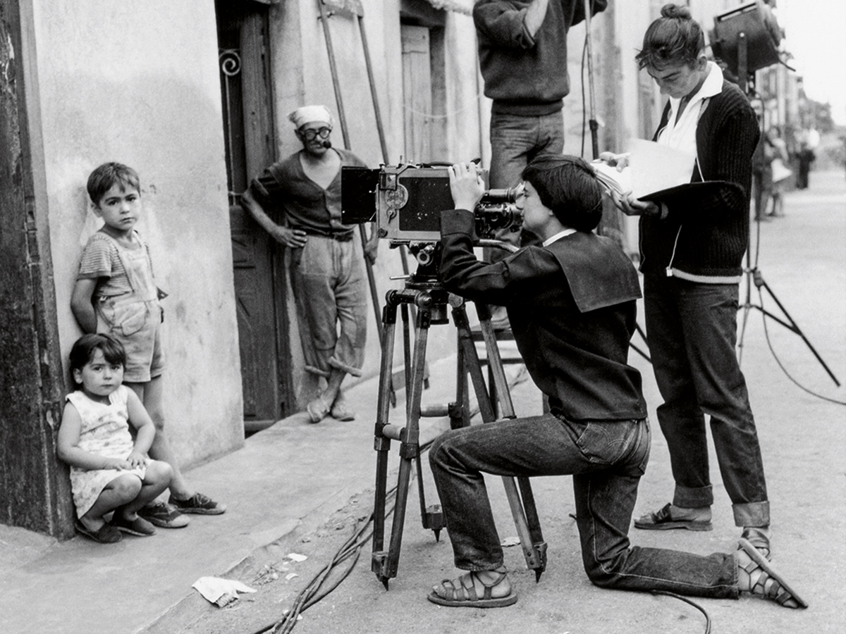 In this black-and-white photo, Agnès Varda is kneeling in order to look through the viewfinder of a large camera. It's focused on a little boy and girl leaning against the wall of a village dwelling. The boy, who is standing, is wearing overall shorts; the girl, who is sitting on her haunches, is younger than him and wears a sleeveless dress. A local man is watching the proceedings. A young woman at Agnès's shoulder, clearly an assistant, appears to be reviewing the script. A man is adjusting the lighting. Everything above his shoulders is cut off.