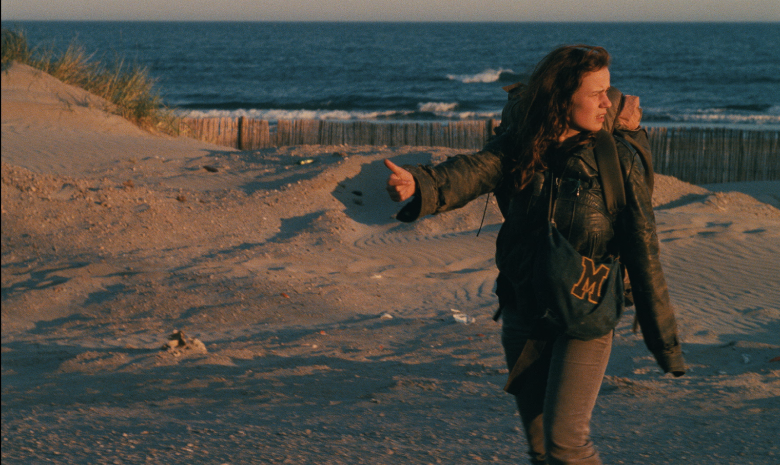 In this color still from Agnès Varda's 1985 film, Vagabond, a young woman (played by Sandrine Bonnaire) is hitchhiking at a beach. She's standing right of center in the foreground, her right arm stretched out and thumb pointing. She is looking back, presumably at a road (one that we do not see) to scan it for a potential ride. Her hair is long and tangled. She's wearing grimy jeans and a dirty leather jacket that looks too big. Along with having a heavy rucksack on her back, she's carrying a cloth bag with a stenciled "M" outlined in orange thread over her shoulder. There are tufts of sea grass and the ocean in the background.