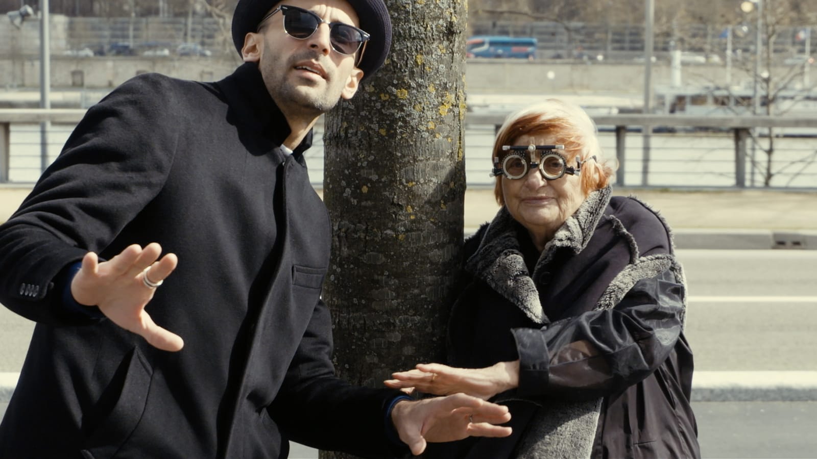 JR (left) stands in dark glasses next to Agnès Varda (right), who is wearing the kind of optical lenses doctors use to assess acuity during routine eye exams.