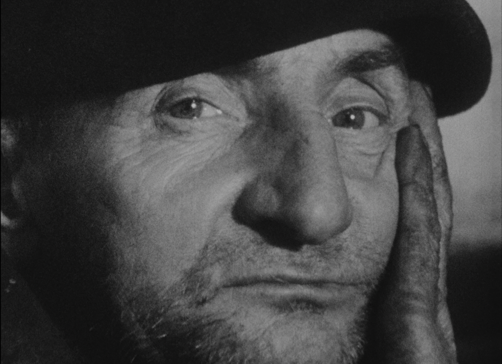 A still from Agnès Varda's black-and-white 1958 film, L’Opéra-Mouffe. A close-up of an aging man with stubble in a beret. He looks into the camera, a serious expression on his face.