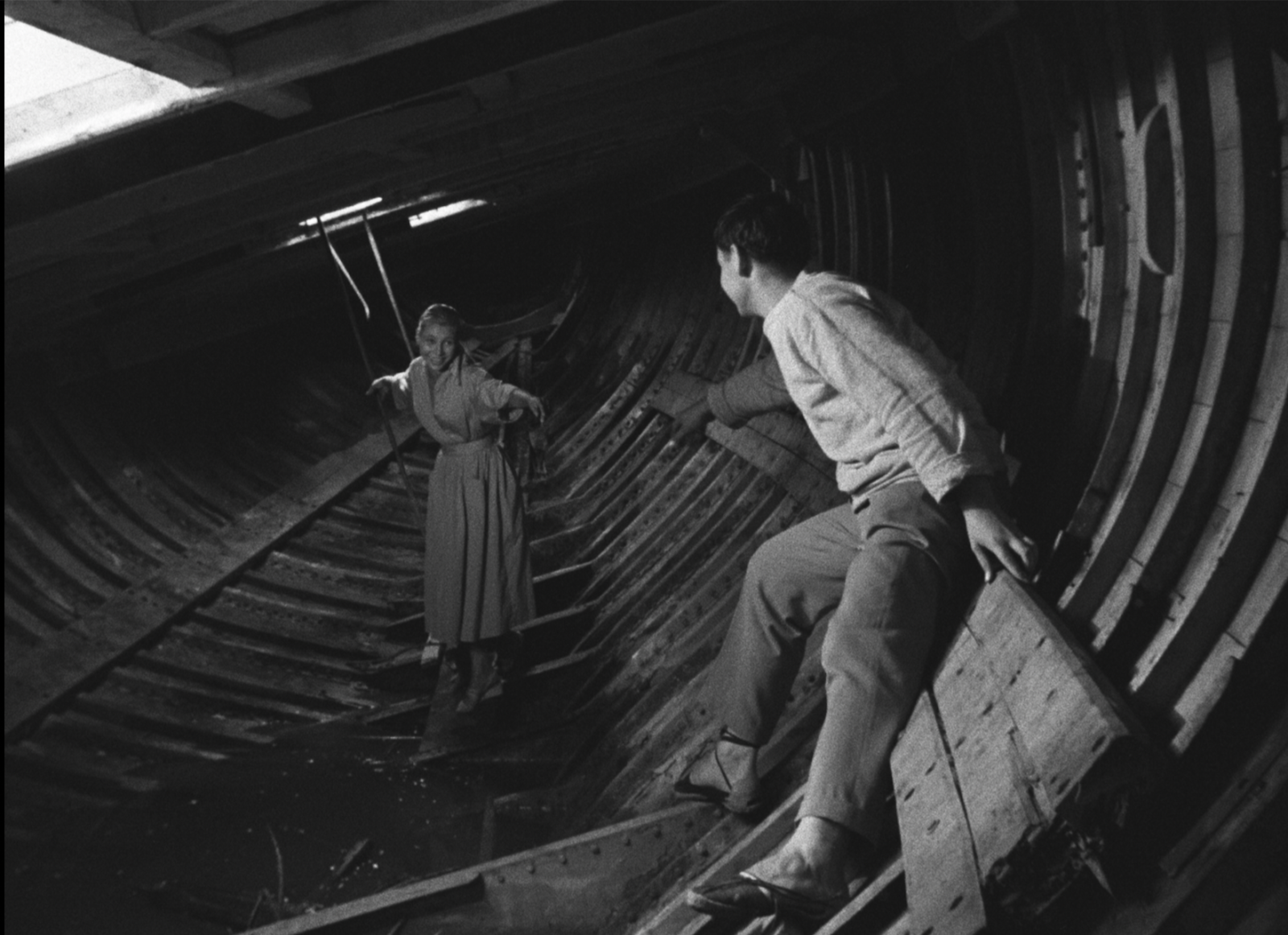 In a still from Agnès Varda’s 1955 black-and-white film La Pointe Courte, a young couple is inside the beached remains of an old hull of a ship. The woman balances herself on the sloping bottom. The man has anchored himself against the side wall and is reaching out to her.