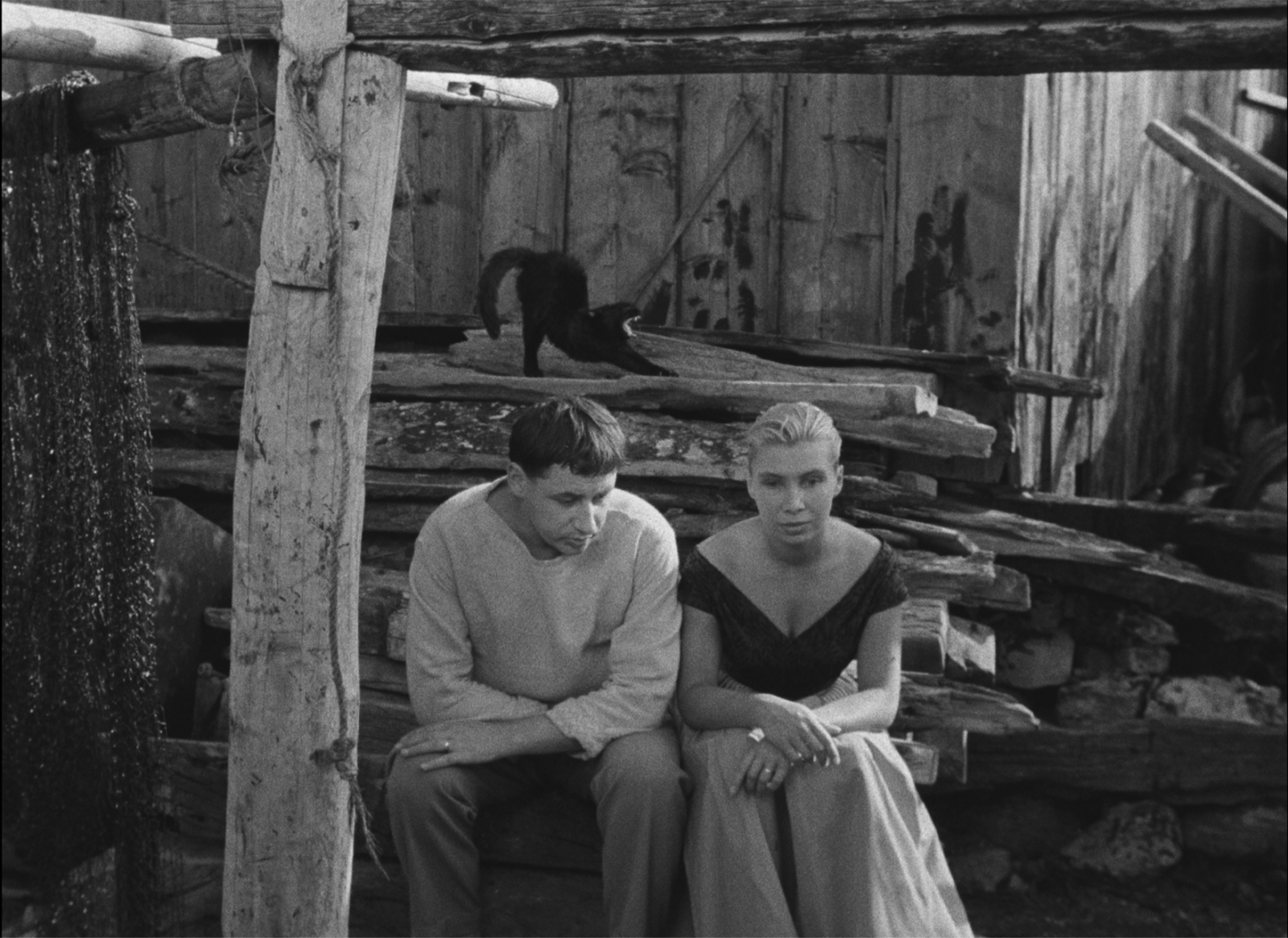 A still from Agnès Varda's 1955 black-and-white film, La Pointe Courte. A young couple, played by actors Philippe Noiret and Silvia Monfort, sit next to one another in front of a shed with a pile of wood and fishing nets. Their arms crossed in front of themselves, he directs his gaze downwards, and she stares unseeing at a point just ahead. A black cat hisses on the woodpile behind them.