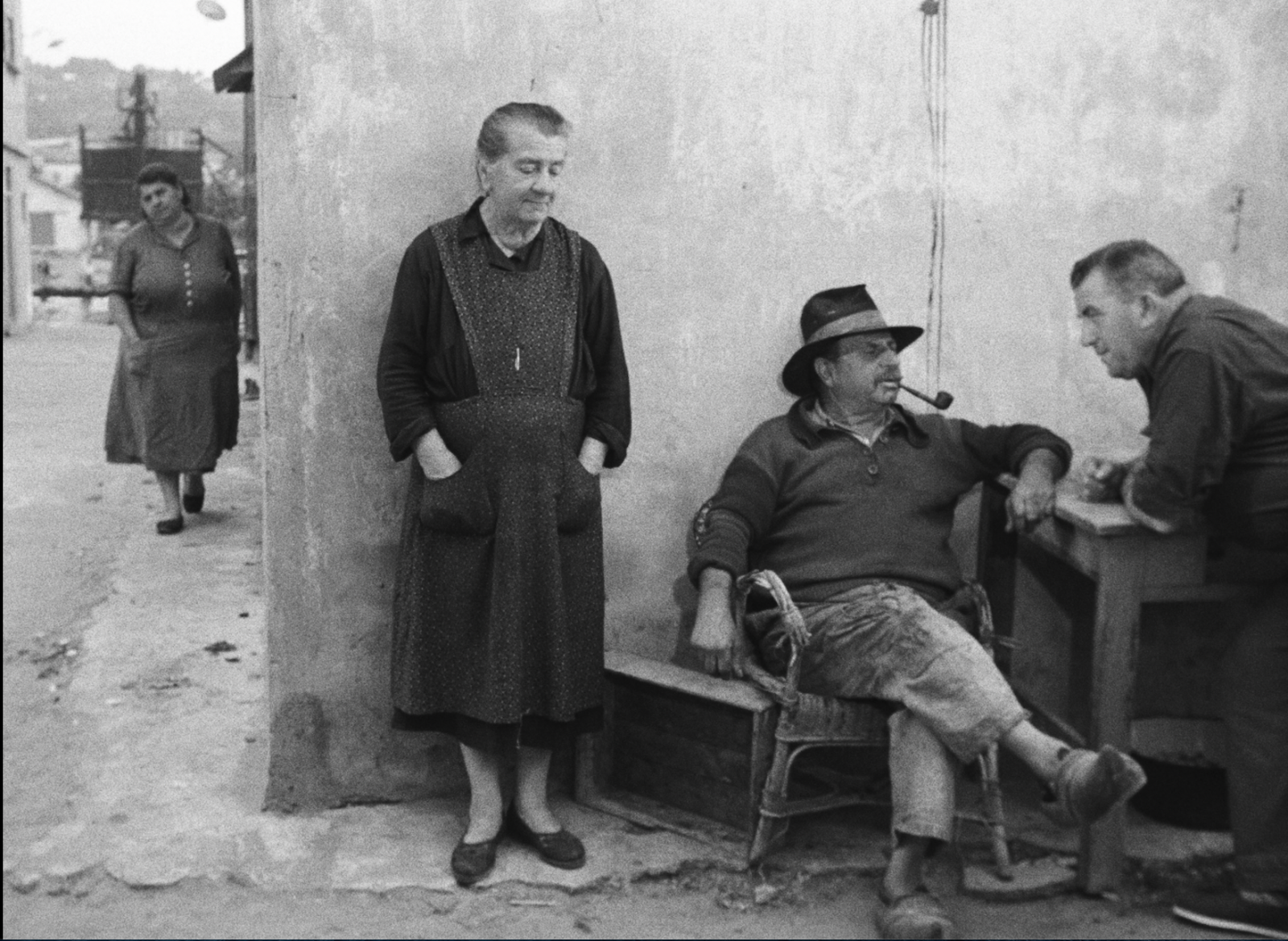 A still from Agnès Varda's 1955 black-and-white film, La Pointe Courte. Four people are outside near a house in a French village. On the right-hand side of the photo, two men chat. One leans in from the right edge of the frame, his arms resting on a table. He's only partially visible. The other sits on a worn wicker chair, his legs crossed. He has a mustache, is smoking a pipe, and wears a hat, grimy pants, and wooden clogs. An older woman in a smock stands to his left, listening. A woman in the background walks toward them.