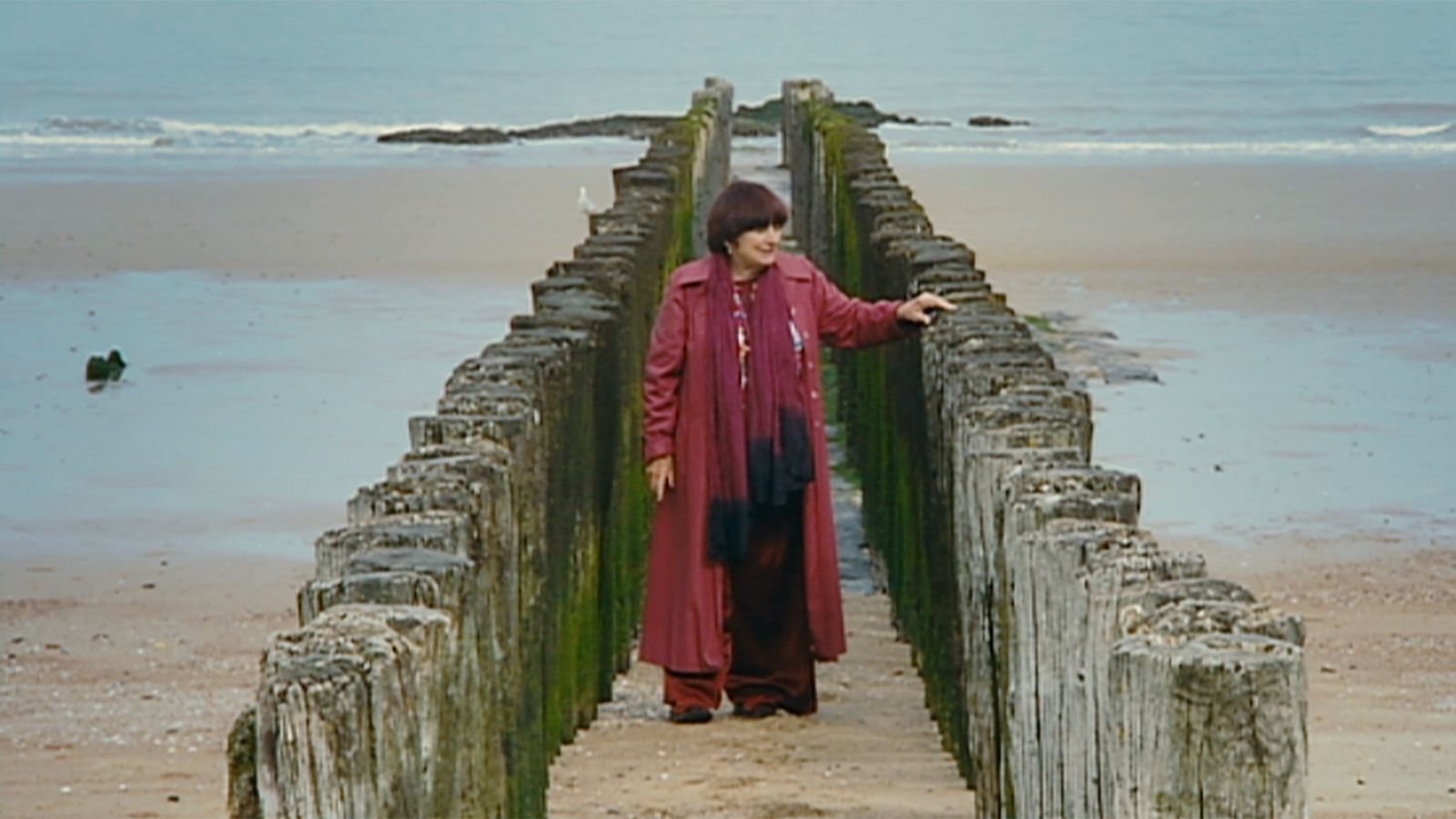 A still from Agnès Varda's 2008 film, The Beaches of Agnès. Wearing a long red coat and scarf, Agnès Varda stands on a causeway.