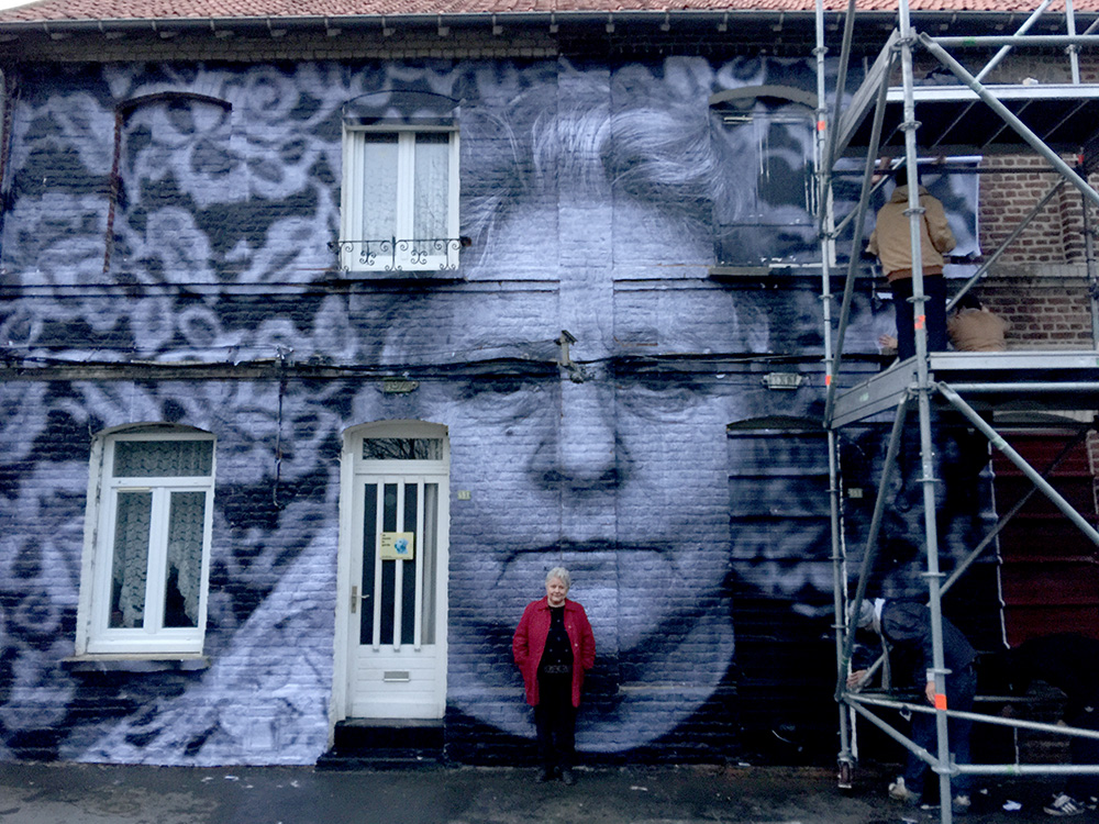 A woman in a red jacket with short white hair stands in front of a gigantic photographic portrait of herself that's been pasted to the brick facade of a house. On the right side of the picture, you can see the scaffolding that was used to paste up the photo, along with two workers.