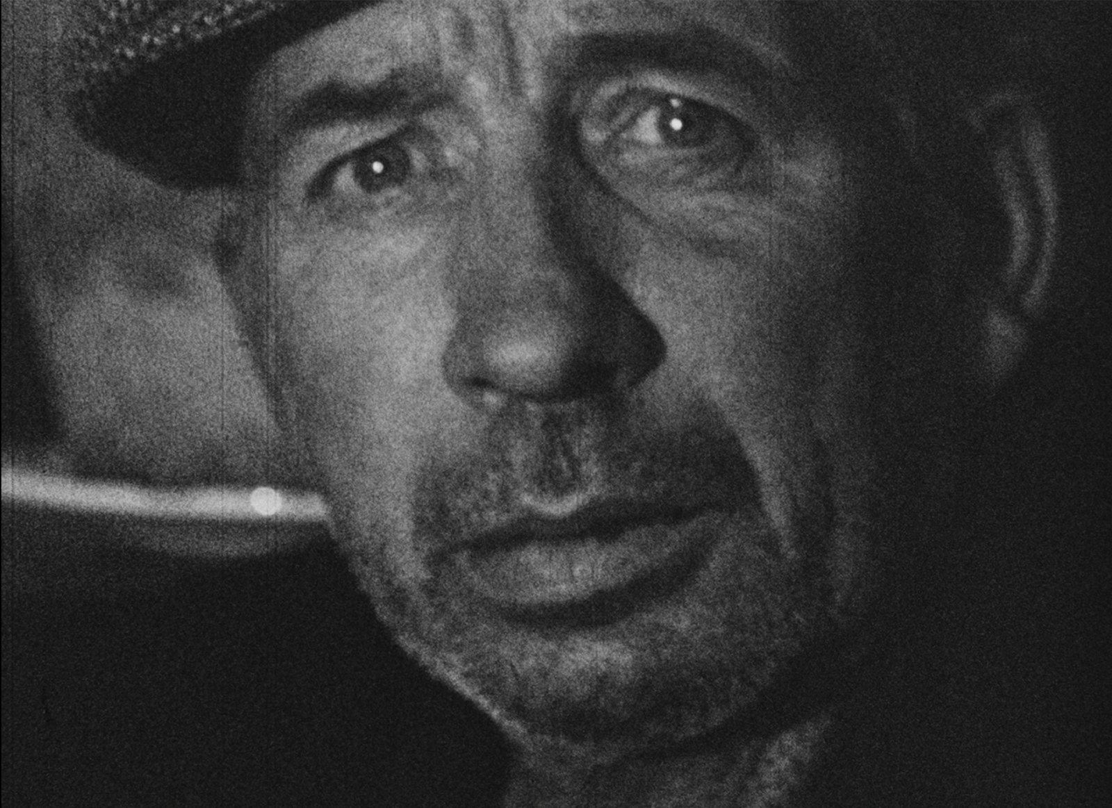 A still from Agnès Varda’s 1958 black-and-white film L’Opéra-Mouffe. Portrait of an older man with an unshaven face, head tipped slightly to the left, deep wrinkles and full lips. He looks directly at the viewer with pursed brows that speak of concern or worry. The image is cropped slightly above his brow to reveal a tweed driver’s cap tipped to one side.