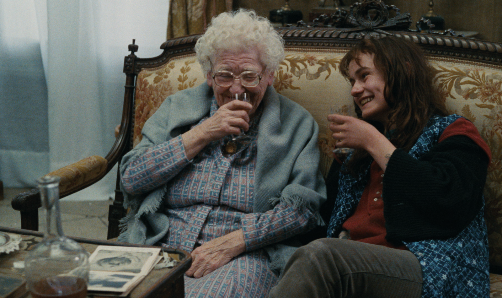 In a still from Agnès Varda’s 1985 film, Vagabond, a young woman (right) drinks cognac with an old woman (left) in a sitting room with an antique settee. They look like they’re having a great time. There are sheer curtains at the window and an album of old photos on the coffee table, next to a clear glass decanter. The old woman wears a day dress with a matching shawl. The young woman is wearing a mishmash of what looks like found clothing: dark jeans, a duotone red-and-black top with bell-shaped sleeves, and a blue patterned vest.