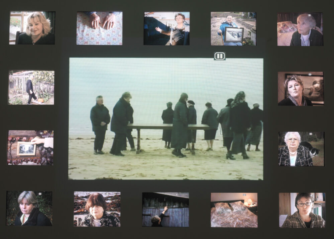 In the center of this video installation, about a dozen women dressed in black walk around a table on a beach. Fourteen smaller video screens surround this central video. On most of the smaller screens, you see headshots of women speaking.