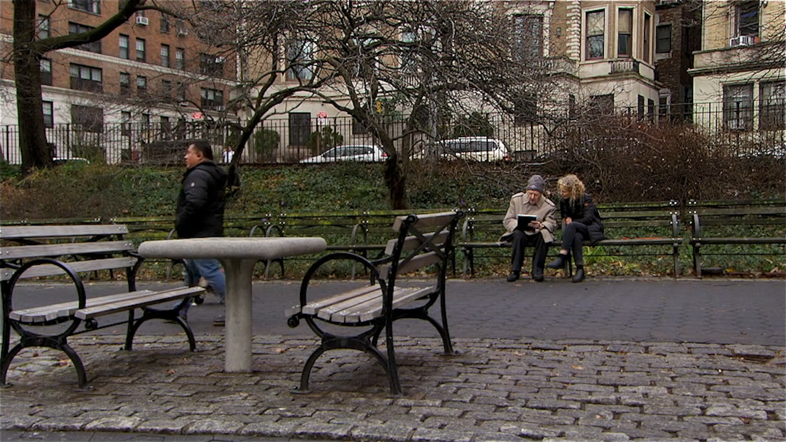 In a still from the film, "Serge Hollerbach: A Russian Painter in New York," Serge Hollerbach and Olga Nenazhivina sit together on a bench in Riverside Park. Serge is wearing a beige coat and cap and is drawing in his sketchbook. Olga, who has curly blonde hair, is leaning towards him to see what he is doing. A pedestrian in a puffy jacket with clippered hair has just passed by in front of them.
