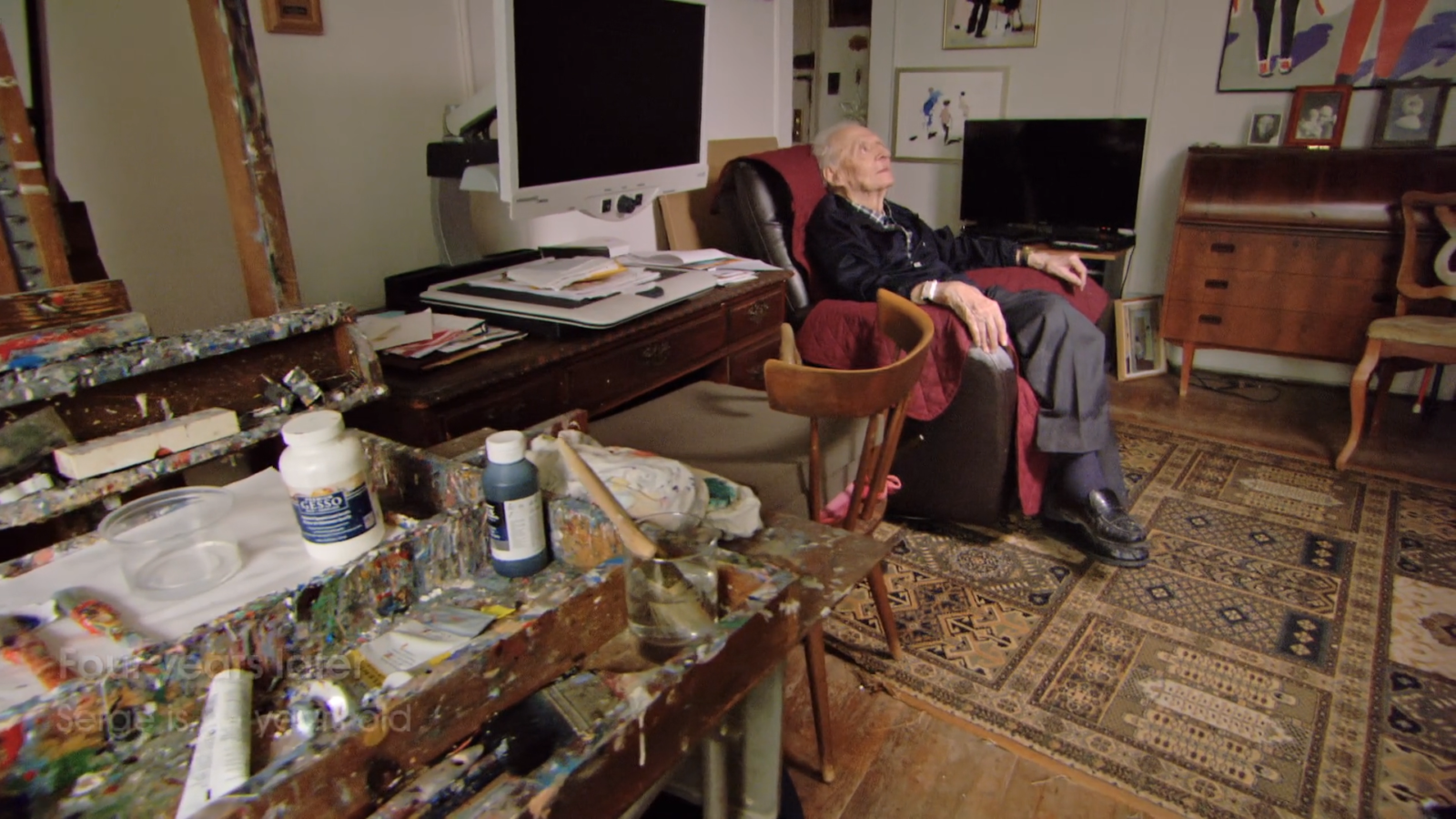 98-year-old artist Serge Hollerbach rests in a recliner in his studio. He is looking up. His palette and easel are in the foreground, crusted with paint from decades of use. There's a desk with a screen reader off to the side and a patterned carpet on the scuffed wood floor. Photographs from his childhood rest on a credenza on the far wall, which is hung with many of Hollerbach's more abstract, post-macular paintings.