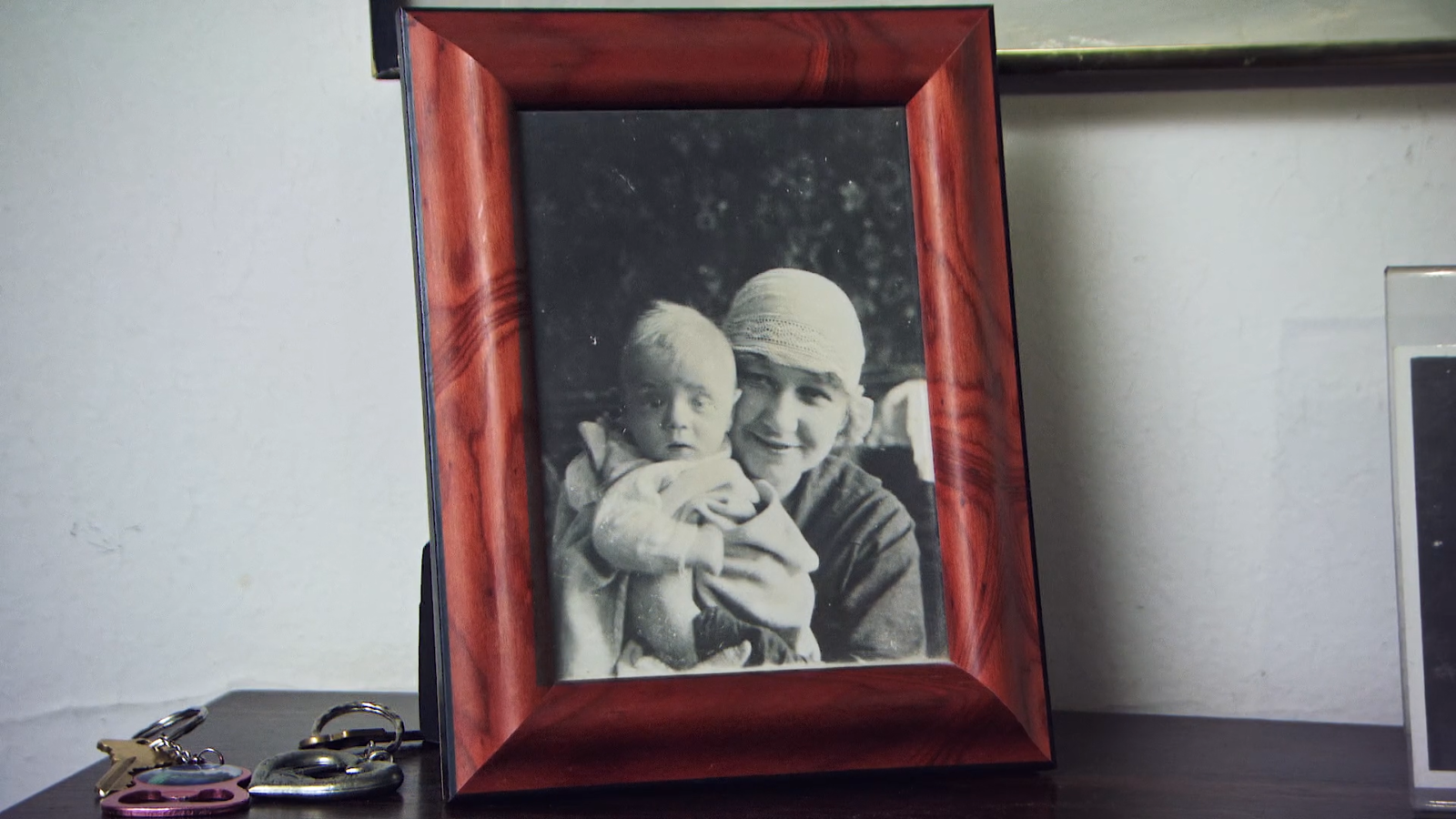 A black-and-white photograph of Serge Hollerbach as a baby with his mother in the early 1920s in Pushkin, Russia. His eyes look startled as he gazes into the camera, and his mother, who is holding him, is wearing a woven hat. The picture's in a red wooden frame on top of a credenza. Next to it are two sets of keys.